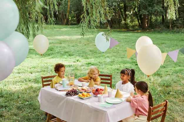 Gruppo di bambini al tavolo da picnic all'aperto decorato con palloncini per la festa di compleanno nello spazio estivo della copia
