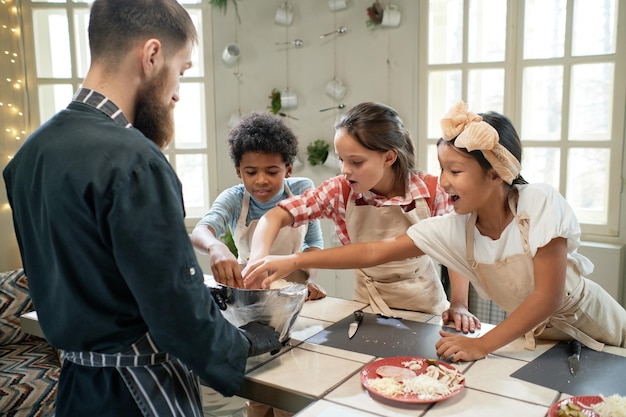 Gruppo di bambini che impastano l'impasto in una ciotola e aiutano lo chef a cucinare durante la lezione di cucina