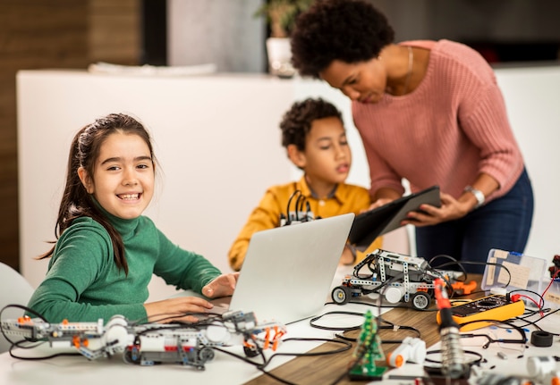 Gruppo di bambini felici con il loro insegnante di scienze femminile afroamericano con laptop che programmano giocattoli elettrici e robot in aula di robotica