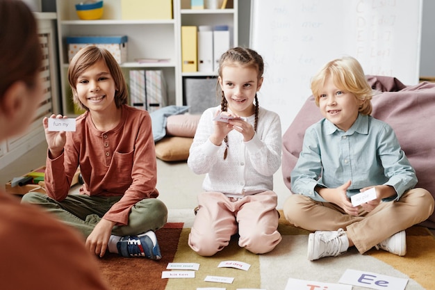 Gruppo di bambini felici seduti sul pavimento e che giocano in inglese insieme all'insegnante in th...