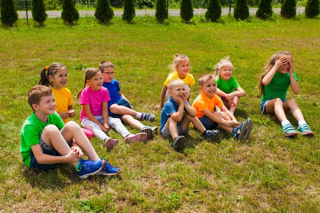 Gruppo di bambini in età prescolare felici che indossano magliette multicolori seduti insieme e ridono sull'erba nel campo estivo.