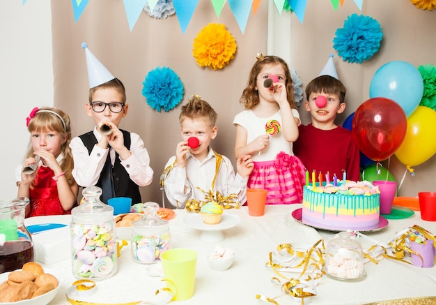 Gruppo di bambini sorridenti che giocano alla festa di compleanno nella stanza decorata. Bambini felici che soffiano nei tubi alla festa di compleanno