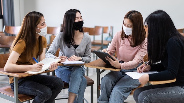 Gruppo di diversi studenti internazionali che indossano maschere protettive e parlano, discutono di progetti, seduti alla scrivania in classe all'università