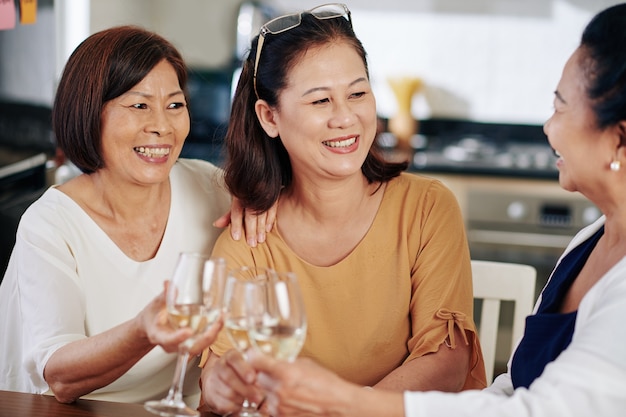 Gruppo di donne anziane asiatiche allegre che bevono vino bianco al tavolo della cucina e parlano di problemi di salute e familiari