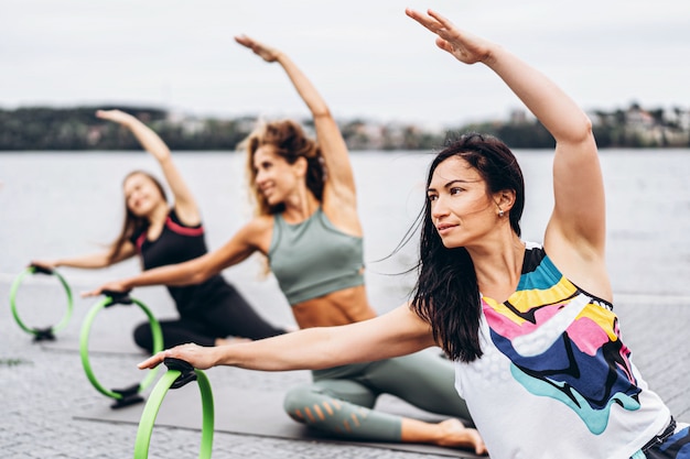 gruppo di donne sportive facendo esercizi di stretching con un cerchio sportivo speciale sulla strada vicino all'acqua.