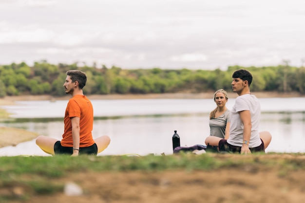 Gruppo di due uomini e una donna che si allungano mentre fanno yoga in un parco vicino a un lago