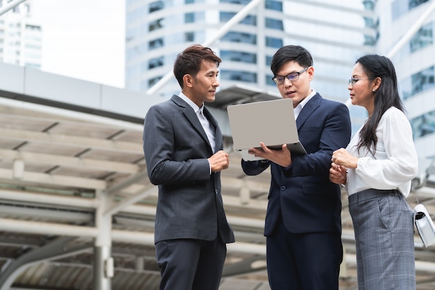 Gruppo di gente di affari asiatica che lavora e che discute qualcosa di positivo con il suo collega maturo e che utilizza un computer portatile ad all'aperto nella capitale