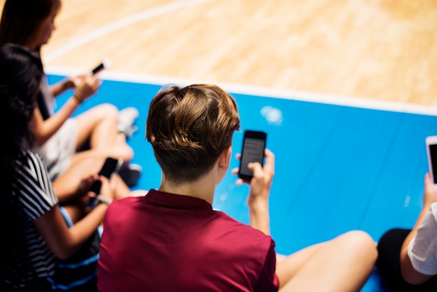 Gruppo di giovani amici dell&#39;adolescente su un campo da pallacanestro che si distende e che per mezzo dello smartphone