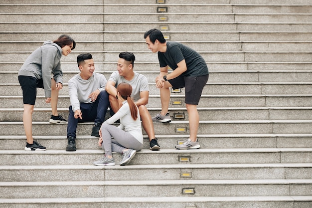 Gruppo di giovani asiatici felici che riposano sui gradini dopo aver corso insieme e aver discusso della maratona