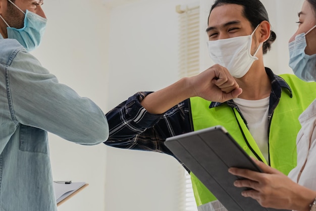 Gruppo di giovani caucasici sorridenti, colleghi o appaltatori asiatici, ingegnere che indossa maschere per il viso e urta i gomiti per proteggersi sul posto di lavoro mentre il coronavirus è pandemico.Saluto nel concetto di Covid,