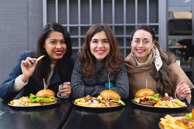 Gruppo di giovani donne attraenti multirazziali che esaminano la macchina fotografica. Femmine sedute a tavola a mangiare hamburger.