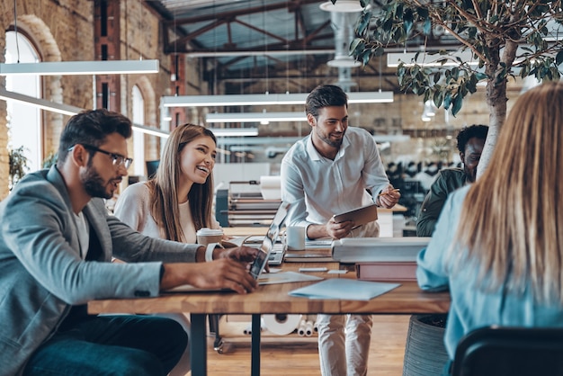 Gruppo di giovani moderni in abbigliamento casual intelligente che comunicano e utilizzano tecnologie moderne mentre lavorano in ufficio