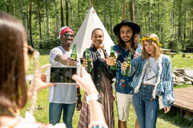 Gruppo di giovani multietnici positivi in abiti hippie in posa con bottiglie di birra per foto al festival di campagna