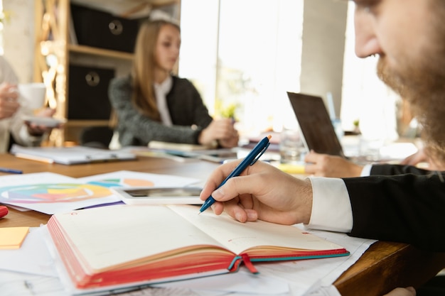 Gruppo di giovani professionisti che hanno una riunione. Diversi gruppi di colleghi discutono di nuove decisioni, piani, strategie. Luogo di lavoro creativo, affari, finanza, lavoro di squadra. Primo piano dell'uomo di scrittura.