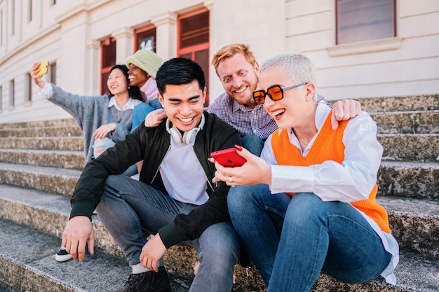 Gruppo di giovani seduti all'aperto a guardare i socialmedia nello smartphone Felice multietnico h