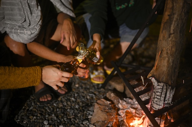 Gruppo di giovani seduti vicino a un falò acceso e bevendo della birra durante il campeggio durante le vacanze del fine settimana