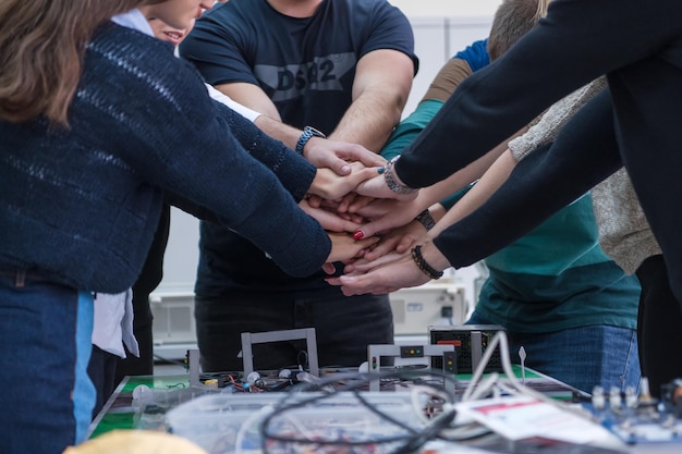 Gruppo di giovani studenti in aula di elettronica che celebrano con successo il progetto finito tenendo le mani insieme, concetto di istruzione e tecnologia