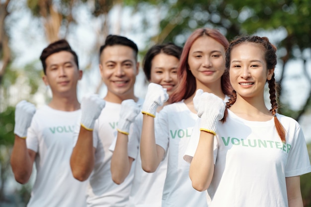 Gruppo di giovani volontari che indossano guanti protettivi quando sono in piedi uno dietro l'altro e sorridono alla telecamera facendo pompa a pugno