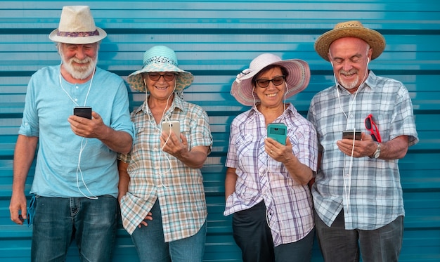 Gruppo di persone anziane in amicizia in piedi contro un muro sorridenti e guardando il telefono ascoltando con gli auricolari. Facce allegre e rilassate per il pensionato