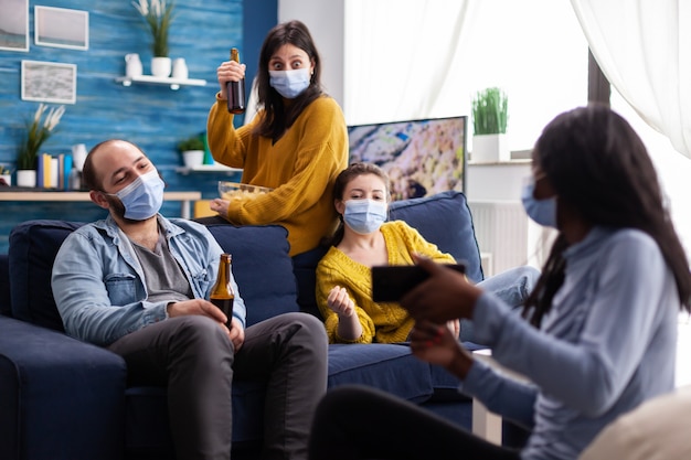 Gruppo di persone diverse che guardano video su smartphone nel soggiorno di casa mantenendo le distanze sociali indossando la maschera facciale come prevenzione durante l'epidemia globale di coronavirus.