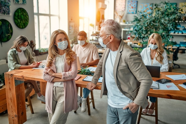 Gruppo di persone multirazziali che lavorano in un ufficio moderno durante il coronavirus. Giovani uomini d'affari e capo senior stanno lavorando insieme indossando maschere protettive per il viso. Misure preventive durante il Covid-19