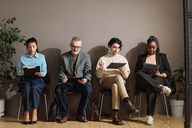 Gruppo di persone sedute su sedie in corridoio e che compilano moduli o scrivono curriculum prima del colloquio di lavoro