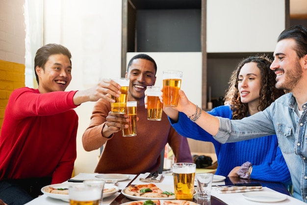 Gruppo di quattro amici multietnici che mangiano pizza in un ristorante e fanno il tifo con bicchieri di birra