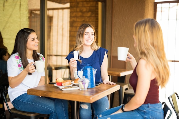 Gruppo di ragazze che bevono caffè e spettegolano al ristorante, con una donna in possesso di carta di credito per il pagamento.