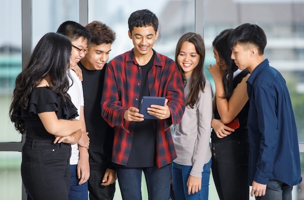 Gruppo di sette giovani adolescenti maschi e femmine in abiti casual in piedi e guardando lo schermo del tablet con un sorriso attraente. Studente universitario che utilizza tablet per chiamare video online con un amico