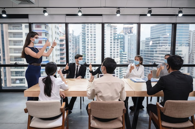 Gruppo di uomini d'affari multietnici che indossa la maschera per il viso che mostra i pollici in su e concordano di un accordo commerciale durante la riunione nel nuovo ufficio normale