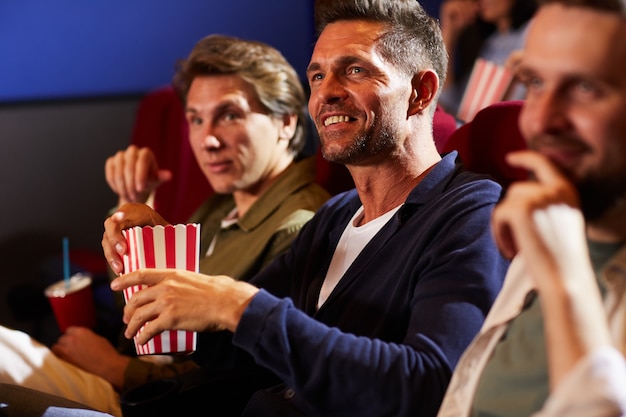 Gruppo di uomini sorridenti che guardano film al cinema e mangiano popcorn seduti in fila sui sedili rossi, copia dello spazio
