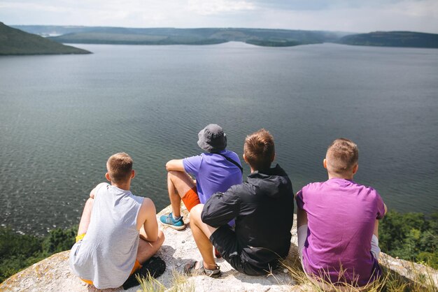 Gruppo di viaggiatori hipster seduti in cima alla montagna rocciosa e godendo di una splendida vista sul fiume Giovani amici esploratori che si rilassano e viaggiano Atmosfera tranquilla momento Copia spazio