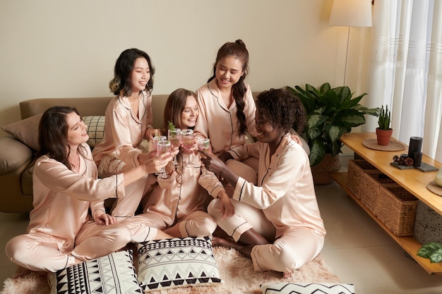 Gruppo eterogeneo di amici in pigiama di seta seduti sul pavimento in camera da letto e brindando con margarita