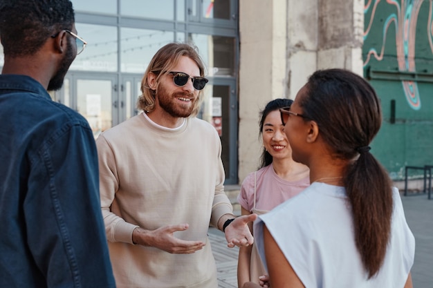 Gruppo eterogeneo di giovani contemporanei che chiacchierano all'aperto in un ambiente urbano, concentrarsi sull'uomo sorridente che indossa occhiali da sole