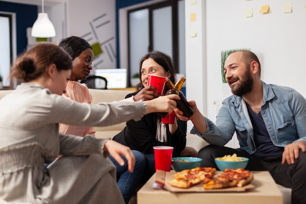 Gruppo eterogeneo di lavoratori che si divertono dopo il lavoro alla riunione dell'ufficio. Amici allegri salutano bottiglie e bicchieri di birra per celebrare la pausa dagli affari. Persone multietniche che sorridono