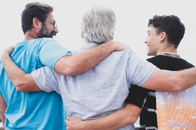 Gruppo familiare multigenerazionale in piedi all'aperto in riva al mare, abbracciati e sorridenti. Nonno, figlio e nipote adolescente.