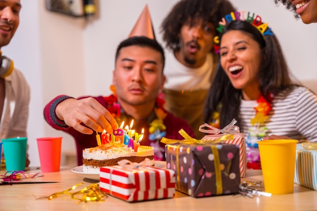 Gruppo multietnico di amici a una festa di compleanno sul divano di casa con una torta e regali Uomo cinese che accende le candele