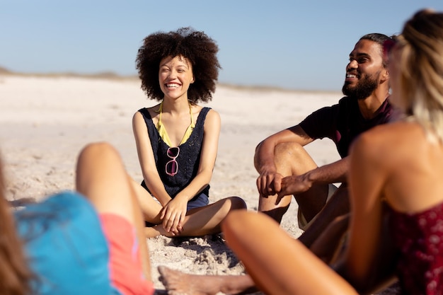 Gruppo multietnico di amici adulti maschi e femmine che indossano pantaloncini e magliette, seduti su una spiaggia assolata, parlano e ridono insieme, con cielo blu e mare sullo sfondo