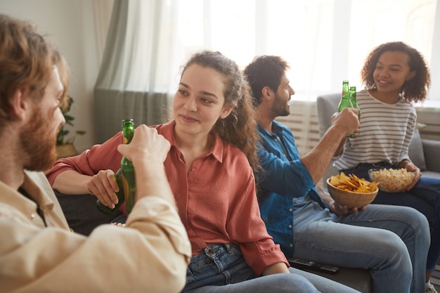 Gruppo multietnico di amici tintinnio di bottiglie di birra mentre si guarda la TV insieme seduti su un comodo divano a casa, concentrarsi sulla coppia giovane in primo piano