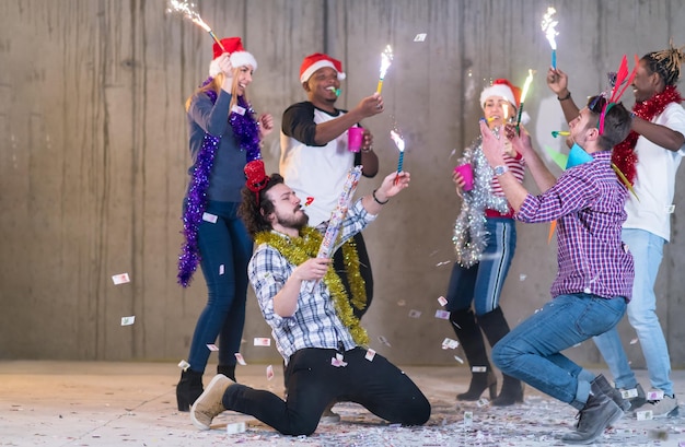 gruppo multietnico di giovani uomini d'affari casuali felici che ballano e fanno festa di coriandoli mentre festeggiano la vigilia di Capodanno davanti al muro di cemento nel nuovo ufficio di avvio