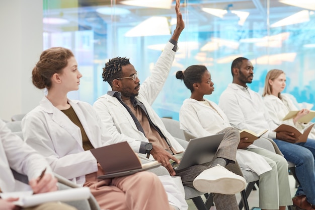 Gruppo multietnico di persone che indossano camici da laboratorio seduti in fila tra il pubblico al seminario medico, concentrarsi sull'uomo afroamericano che alza la mano per fare domande, copia spazio