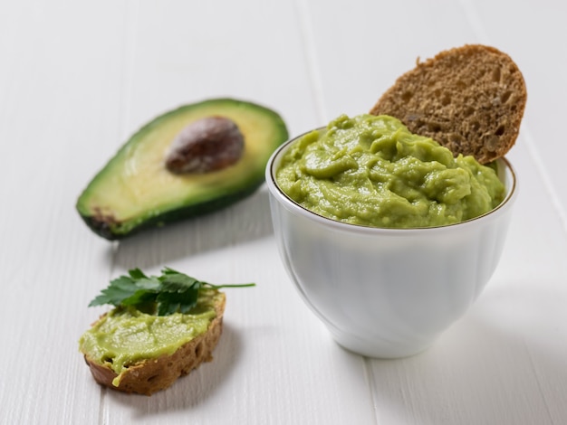 Guacamole in una tazza bianca con fette sottili di pane su un tavolo di legno bianco. Dieta vegetariana cibo messicano avocado. Cibo crudo.