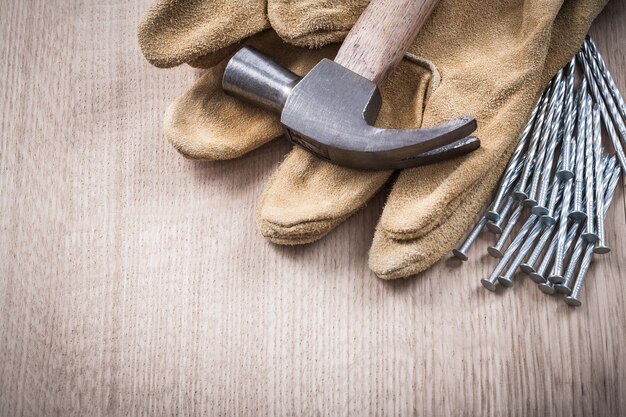 Guanti protettivi in pelle con martello da carpentiere e mucchio di chiodi metallici su tavola di legno versione ravvicinata del concetto di costruzione