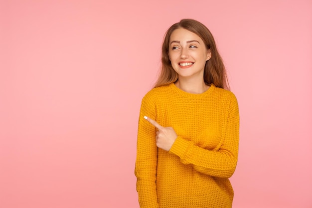 Guarda, attenzione. Ritratto di ragazza allegra attraente zenzero in maglione che punta di lato e sorridente, mostrando lo spazio della copia, posto vuoto per il testo promozionale. girato in studio isolato su sfondo rosa