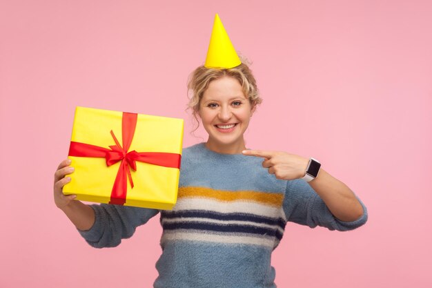 Guarda il mio presente Ritratto di donna felice con buffo cappello a cono sulla testa e in maglione caldo che punta al regalo di vacanza e sorridente con gioia mostrando sorpresa di compleanno in studio indoor girato sfondo rosa