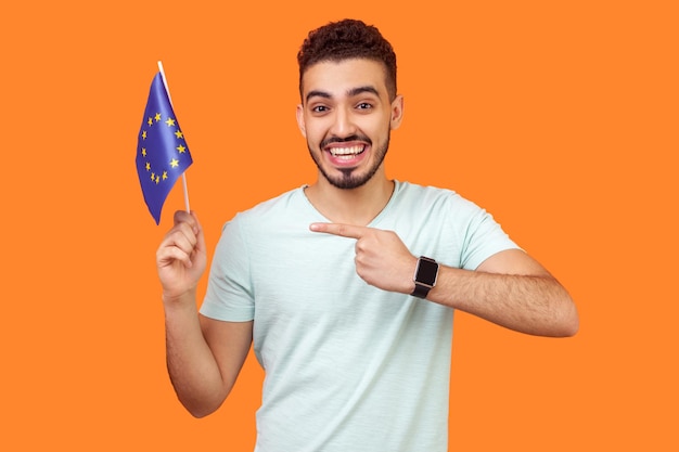 Guarda la bandiera dell'Unione Europea. Ritratto di gioioso eccitato uomo bruna con la barba in t-shirt bianca sorridente e puntando alla bandiera dell'UE, sorridendo alla telecamera. tiro in studio indoor isolato su sfondo arancione