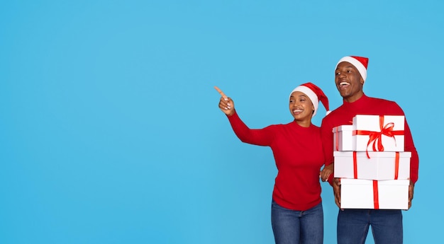 Guarda lì i coniugi afroamericani in cappelli di Babbo Natale che tengono una pila di scatole di regali di Natale e puntano il dito da parte mostrando lo spazio libero per il testo pubblicitario di Natale Panorama su sfondo blu