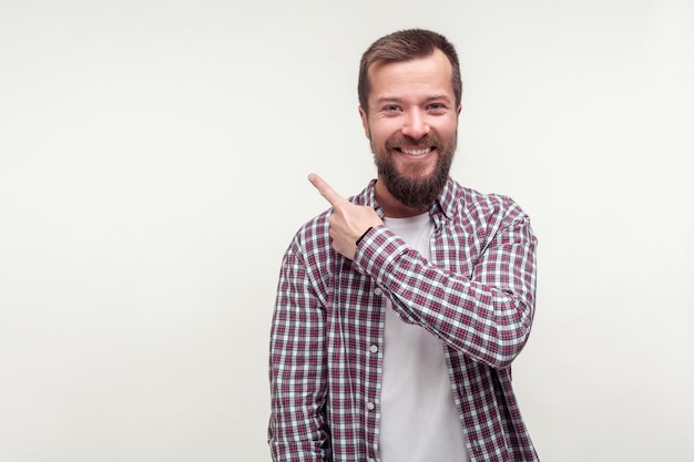 Guarda lì Ritratto di uomo barbuto felice positivo in camicia a quadri casual che punta il dito di lato verso lo spazio vuoto per il posto pubblicitario per il testo in studio indoor girato isolato su sfondo bianco