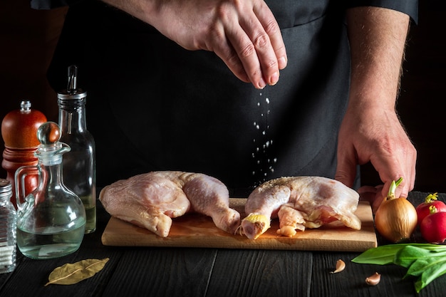 Guarda o lo chef aggiunge sale alle cosce di pollo crude. Preparazione per grigliare con coscia di pollo nella cucina di un ristorante o bar su un tavolo con verdure e spezie