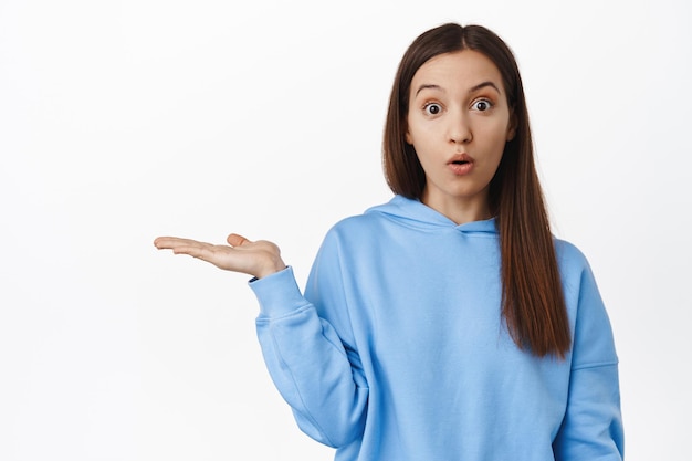 Guarda questo. Giovane donna in felpa con cappuccio che tiene in mano aperta vuota, palmo su sfondo bianco vuoto, ragazza che guarda sorpresa alla telecamera, sfondo bianco